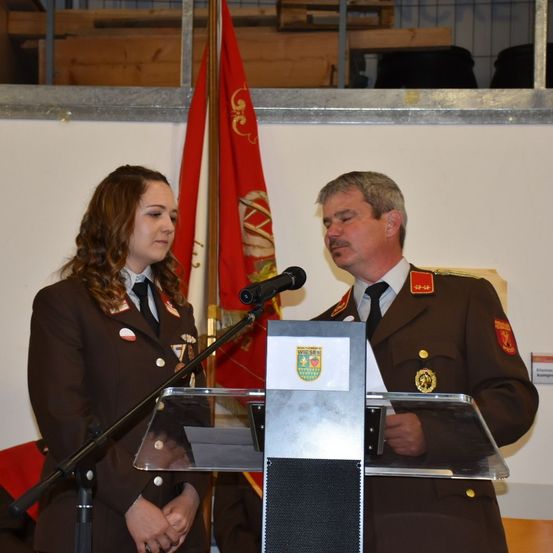 Zwei Personen, ein Mann und eine Frau, in Militäruniformen, stehen vor einem Podium mit einer Flagge dahinter. Der Mann scheint zu sprechen, während die Frau aufmerksam zuhört.