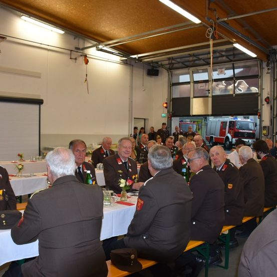 Eine Versammlung von uniformierten Personen in einem Saal, die an Tischen sitzen, mit einem Feuerwehrwagen, der durch das Garagentor sichtbar ist.