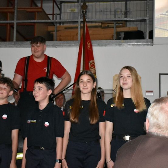 Eine Gruppe von Kindern in Uniform steht auf einer Bühne, mit einer Frau dahinter und einer Flagge. Ein Mann in einem roten Shirt steht hinter ihnen. Im Hintergrund sitzt eine Person, und eine Projektionsleinwand ist sichtbar.