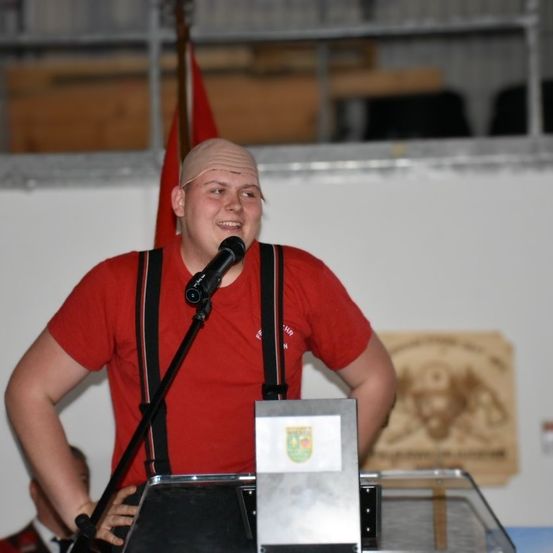 Ein Mann in einem roten T-Shirt und Hosenträger steht an einem Podium, lächelt und hat ein Mikrofon vor sich. Hinter ihm ist eine rote Fahne und ein Schild mit einem Logo zu sehen. Ein weiterer Mensch ist teilweise im Hintergrund sichtbar.