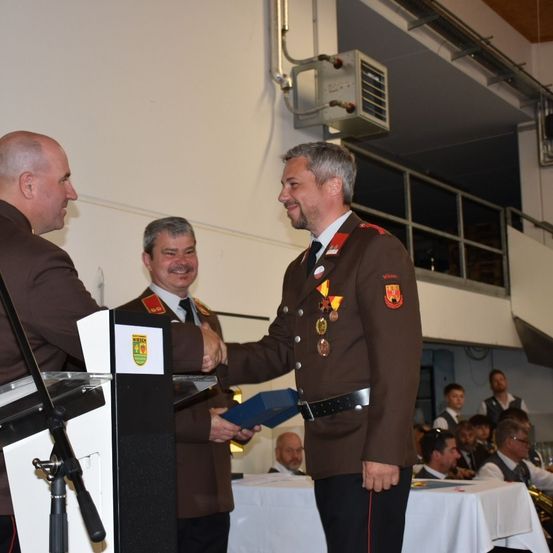 Drei Männer in Uniform stehen vor einem Podium. Einer überreicht dem anderen eine Auszeichnung. Ein Publikum sitzt dahinter.