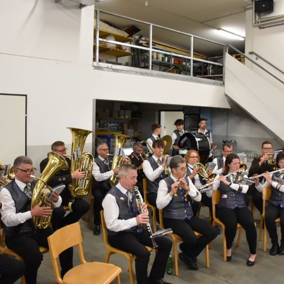 Eine Gruppe von Musikern spielt in einem Lagerhaus, mit Tuba-, Posaunen- und Flötenspielern, die auf Stühlen sitzen. Der Raum hat weiße Wände, eine Treppe und eine Zwischenebene mit Regalen.