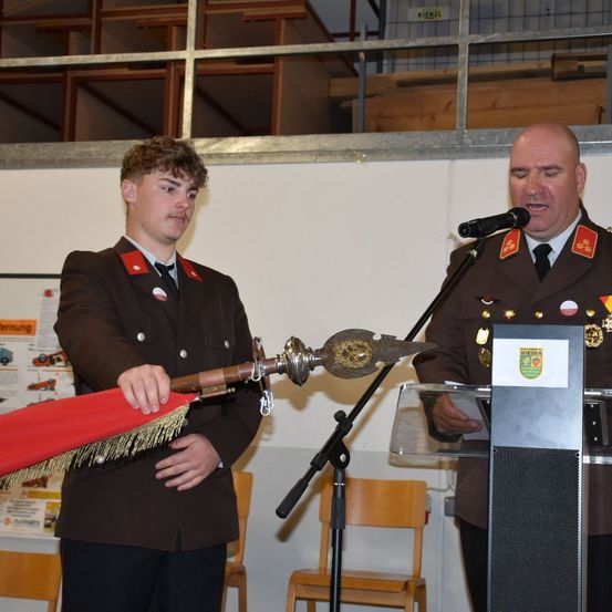 Ein Mann in Uniform steht an einem Rednerpult mit Mikrofon, während ein anderer Mann ein zeremonielles Schwert hält.