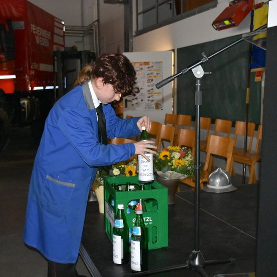 Eine Person in einem blauen Mantel öffnet auf einer Bühne eine Bierflasche, mit einem Mikrofon und einer Tafel im Hintergrund.