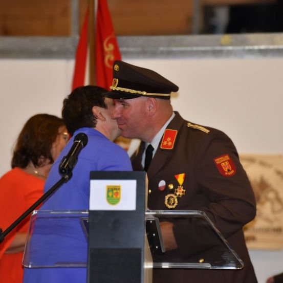 Ein Mann in Uniform küsst eine Frau in einem blauen Kleid am Rednerpult. Vor ihnen steht ein Mikrofon. Dahinter sind eine Flagge und eine Frau in einem orangefarbenen Kleid zu sehen.