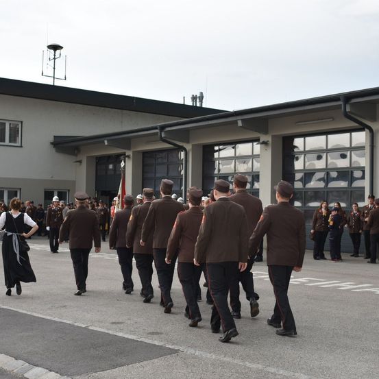 Eine Gruppe von Menschen in braunen Uniformen marschiert an einem Gebäude vorbei. Zuschauer stehen hinter ihnen.
