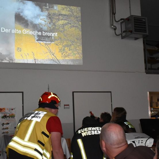 Ein Raum mit einer Projektionsleinwand, die ein Bild eines brennenden Gebäudes mit dem Text 'Der alte Grieche brennt' zeigt. Feuerwehrleute in Uniform sind versammelt, einige sitzen und einige stehen.