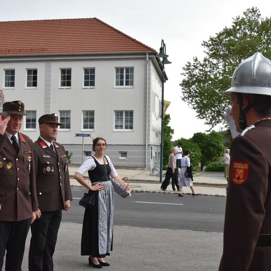Eine Frau in traditioneller Kleidung steht in der Mitte der Straße, flankiert von zwei Männern in Militäruniformen. Hinter ihnen gehen Menschen auf dem Bürgersteig, und ein Gebäude mit roten Dachziegeln ist sichtbar.