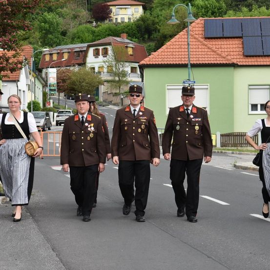 Vier uniformierte Personen gehen auf einer Straße, mit zwei Frauen an der Seite. Gebäude mit Solarpaneelen sind im Hintergrund.