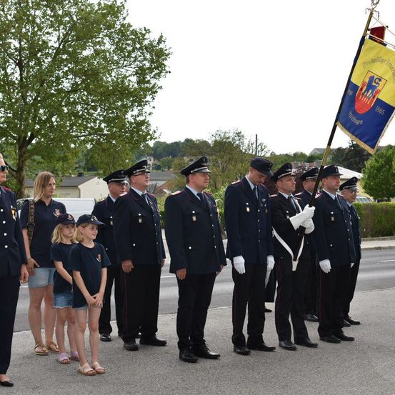 Eine Reihe von uniformierten Personen, möglicherweise Feuerwehrleute, steht in Formation auf einer Straße. Einige halten eine Fahne. In der Nähe stehen einige Frauen und Kinder, und Gebäude sind im Hintergrund zu sehen.