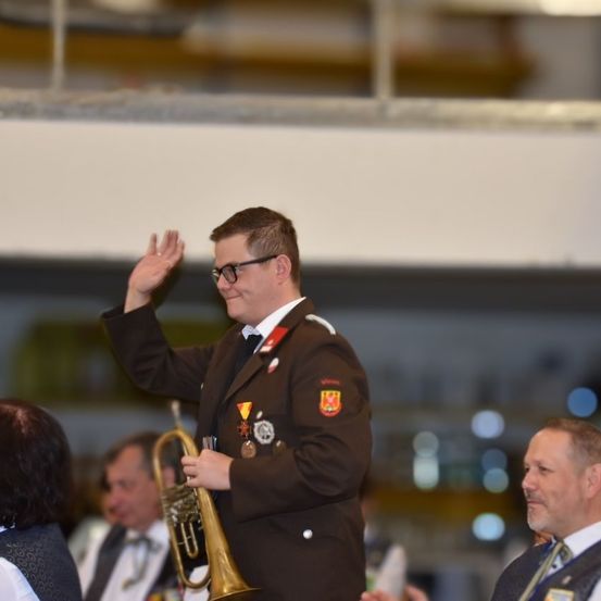 Ein Mann in Uniform winkt, während er eine Trompete hält, lächelt und von anderen bei einer Veranstaltung umgeben ist.