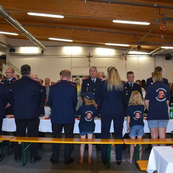 Mehrere uniformierte Personen, einschließlich Kinder, stehen in einem Raum mit Tischen. Der Raum hat eine Holzdecke und Metallrohre. Einige Leute tragen Hüte und Brillen. Ein Kreuz ist an der Wand sichtbar.