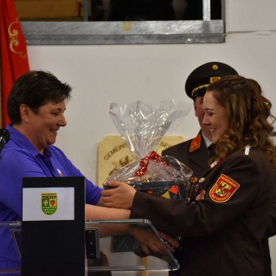 Eine Frau in Uniform überreicht einer lächelnden Frau an einem Podium mit Mikrofon einen Geschenkkorb. Hinter ihnen steht ein Mann in Uniform.