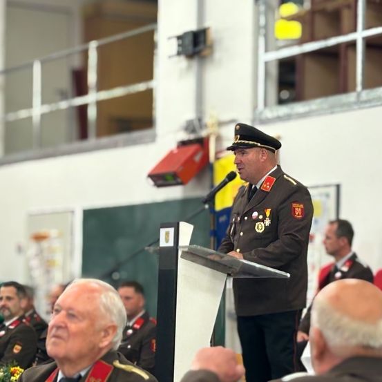 Ein Mann in Uniform spricht an einem Podium vor einem Publikum, mit einem Mikrofon und einer gelben Flagge dahinter.
