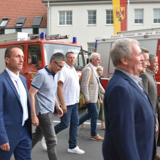Männer in Anzügen und Freizeitkleidung gehen an Feuerwehrwagen in einer Stadt vorbei. Gebäude mit deutschen Flaggen sind im Hintergrund.