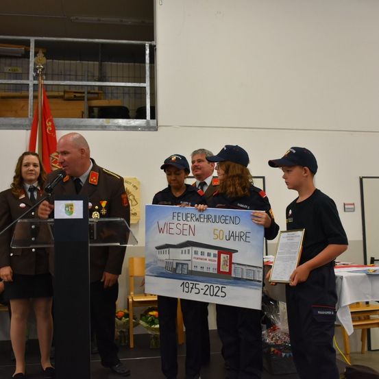 Eine Zeremonie findet statt, bei der ein Mann in Uniform an einem Podium spricht, während eine Frau ein Banner hält, das 'Feuerwehrjugend Wiesen 50 Jahre 1975-2025' liest. Ein junger Junge steht neben ihr und hält eine Plakette.