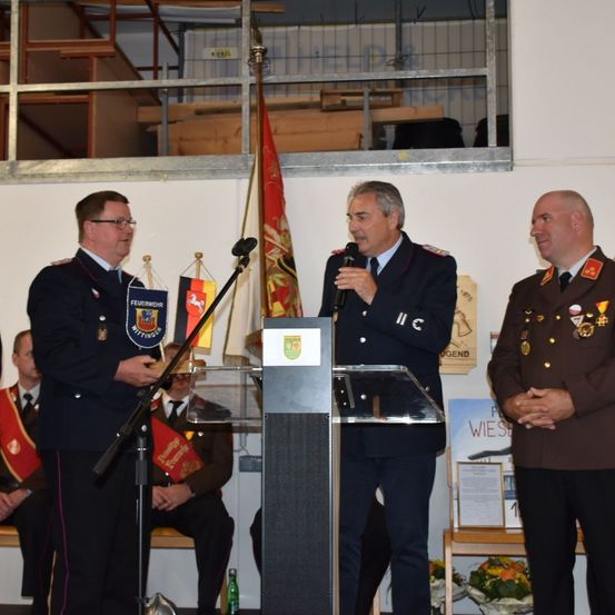 Drei Männer in Militäruniformen stehen vor einem Podium mit Fahnen. Ein Mann spricht in ein Mikrofon. Dahinter sitzen und stehen eine Gruppe von Menschen, die die Zeremonie beobachten.