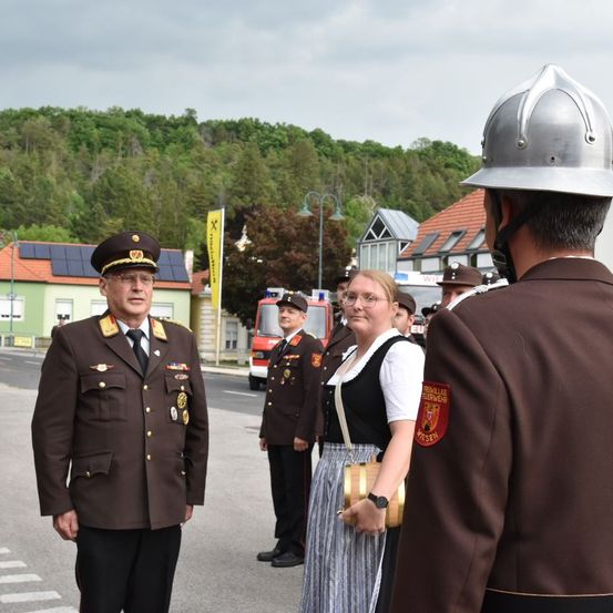 Ein Mann in Uniform steht neben einer Frau in traditioneller Kleidung, beide schauen einen Mann in Feuerwehruniform an.