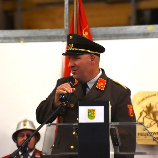 Ein Mann in Uniform steht an einem Rednerpult und spricht in ein Mikrofon. Hinter ihm ist eine Fahne zu sehen. Ein weiterer Mann mit Helm steht in der Nähe.