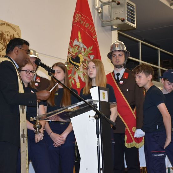 Ein Mann in einem schwarzen Anzug spricht in ein Mikrofon und hält eine feierliche Kerze. Hinter ihm steht eine Gruppe von Menschen, darunter Kinder, vor einem Podium. Ein Kind trägt einen Hut, ein anderes einen Helm. Im Hintergrund befinden sich eine Flagge und Metallgeländer.