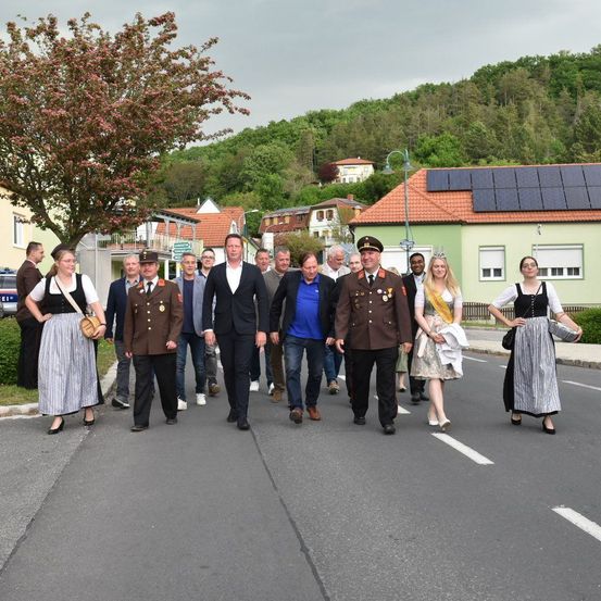 Eine Gruppe von Menschen, darunter ein Mann in Uniform, geht auf einer Straße in einer Stadt. Die Straße hat Häuser mit Solarpanelen auf den Dächern.