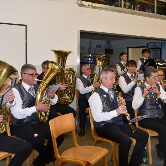Eine Gruppe von Musikern sitzt auf Stühlen und spielt verschiedene Instrumente. Sie tragen formelle Kleidung mit Westen und Krawatten. Einige halten Notenblätter, während andere sich auf ihre Instrumente konzentrieren. Der Raum hat eine weiße Wand und eine Galerie oben.