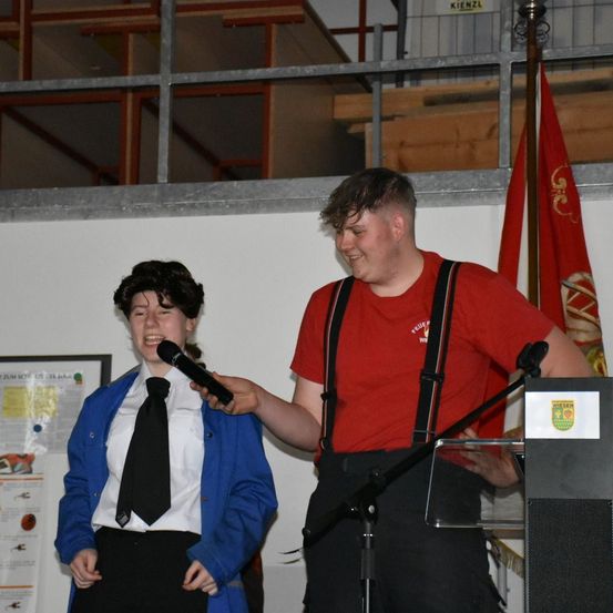 Eine Person in einem roten Shirt mit Hosenträger hält ein Mikrofon an eine lachende Person in einem blauen Mantel und Krawatte. Hinter ihnen ist eine Fahne und ein Podium mit einem Logo.