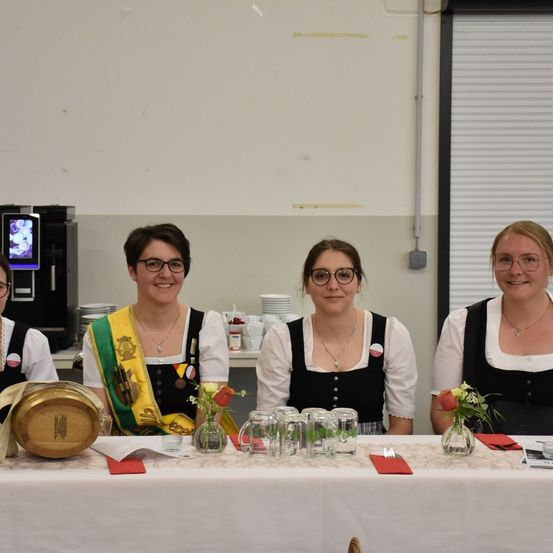 Vier Frauen in traditioneller Kleidung, die Brillen tragen, sitzen hinter einem Tisch mit Blumen, Gläsern und einem Fass. Dahinter befindet sich eine Theke mit Tellern und einer Kaffeemaschine.