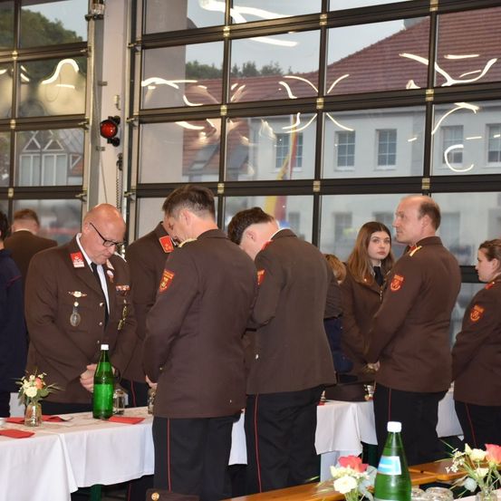 Mehrere Personen in Militäruniformen stehen um einen Tisch herum, wobei ein Mann mit Brille eine Flasche betrachtet. Ein anderer Mann hat einen Blumenstrauß auf dem Tisch. Hinter ihnen sind große Fenster, die ein Gebäude und Bäume zeigen.