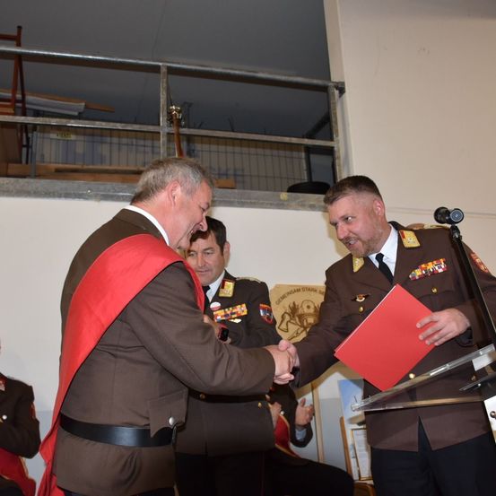 Zwei Männer in Militäruniformen schütteln bei einer Veranstaltung die Hände. Ein Mann hält eine rote Mappe, und der andere trägt einen roten Umhang. Sie stehen vor einem Podium mit einem Mikrofon. Hinter ihnen sieht ein weiterer Mann in Uniform sie an.