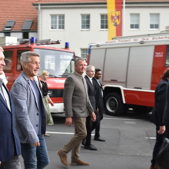 Mehrere Männer in Anzügen gehen auf einer Straße vor einem Feuerwehrauto. Dahinter steht eine Frau in einem grünen Kleid. Das Gebäude im Hintergrund hat eine Flagge oben.