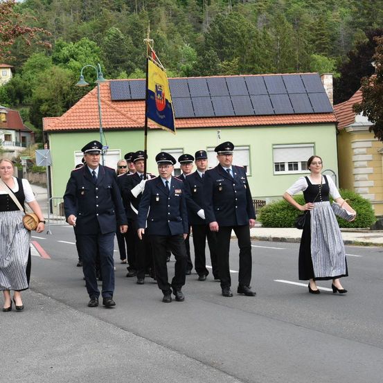 Eine Gruppe von Militäroffizieren und einer Frau in traditioneller Tracht marschiert in einer Parade. Dahinter ist ein Haus mit Solarpanelen auf dem Dach und einem Fahnenmast zu sehen.