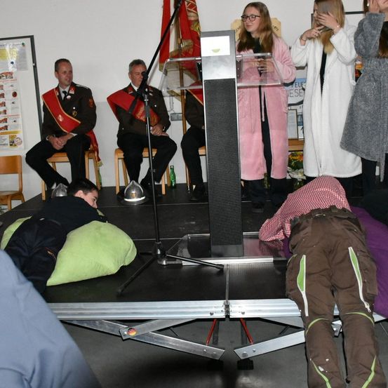 Mehrere Personen sind in einem Raum versammelt; zwei uniformierte Männer sitzen auf Stühlen, eine Frau steht an einem Rednerpult und ein Mann liegt auf dem Boden. Eine weitere Frau in einem rosa Bademantel steht neben dem Rednerpult.