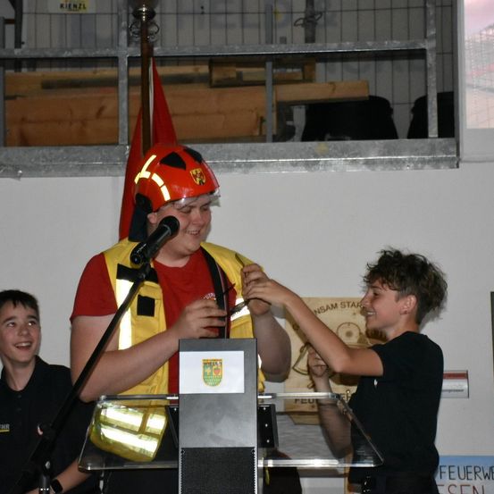 Ein junger Mann in einem roten Helm und einer reflektierenden Weste steht an einem Podium und überreicht einem Jungen in einem schwarzen T-Shirt eine Auszeichnung. Ein weiterer Junge in einem schwarzen T-Shirt steht hinter ihnen. Hinter dem Podium ist eine Fahne sichtbar.