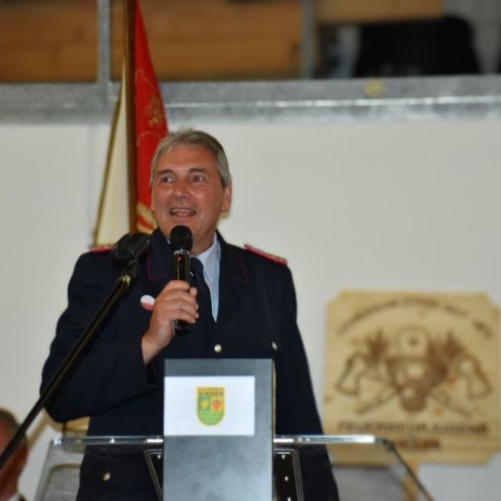 Ein Mann in Uniform steht an einem Rednerpult und spricht in ein Mikrofon. Hinter ihm ist eine Fahne und ein Schild zu sehen. Die Umgebung scheint drinnen zu sein.