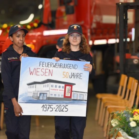 Zwei Frauen in Uniform halten ein Schild mit der Aufschrift 'FEUERWEHRJUGEND WIESEN 50 JAHRE 1975-2025', sie stehen vor einem roten Feuerwehrwagen und einem Blumenstrauß.