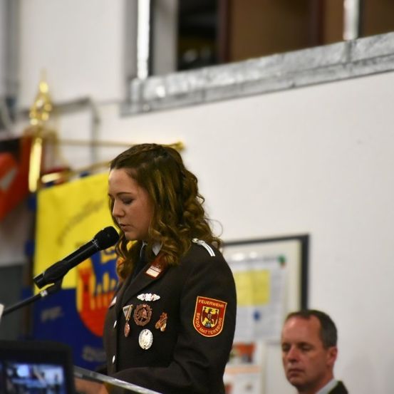 Eine Frau in Uniform steht an einem Rednerpult mit einem Mikrofon. Sie trägt ein Abzeichen und mehrere Medaillen. Hinter ihr befindet sich eine gelbe Fahne und ein Mann im Anzug.