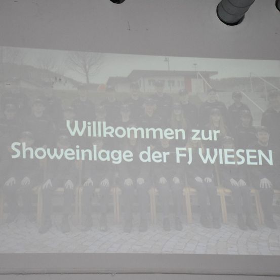 Eine Gruppe von Menschen in Uniformen sitzt auf Bänken, mit einer Willkommensnachricht in Deutsch, die auf einem Bildschirm über ihnen angezeigt wird.