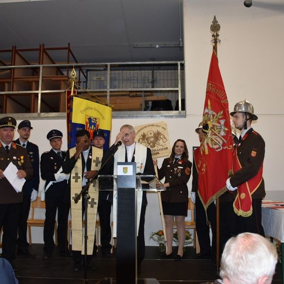 Eine Gruppe uniformierter Personen steht auf der Bühne, darunter ein Mann, der an einem Podium spricht, mit Fahnen hinter ihm. Andere Männer und Frauen stehen neben ihm, und Menschen sitzen im Publikum.