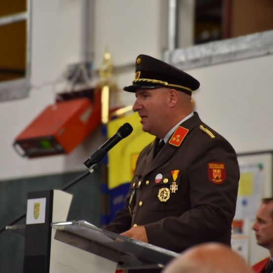 Ein Militäroffizier in Uniform hält eine Rede an einem Rednerpult mit einem Mikrofon vor sich. Hinter ihm befinden sich Fahnen und Dekorationen, und eine weitere Person ist teilweise im Hintergrund zu sehen.