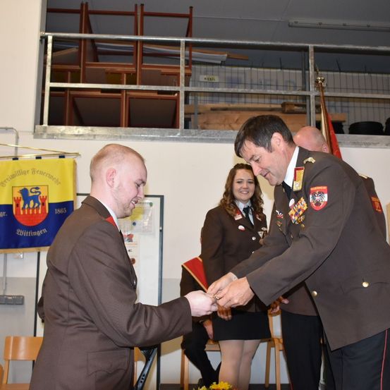 Ein Mann in Uniform überreicht einem anderen Mann eine Medaille. Dahinter lächelt eine Frau. Oben hängt ein Banner.