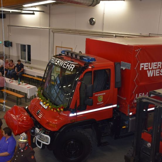 Ein roter Feuerwehrwagen mit der Aufschrift 'Feuerwehr Wiesen' steht in einer Halle. Ein Mann in einer Feuerwehruniform steht neben ihm. Ein Gabelstapler befindet sich rechts. Hinter dem Wagen steht ein Tisch mit Flaschen.