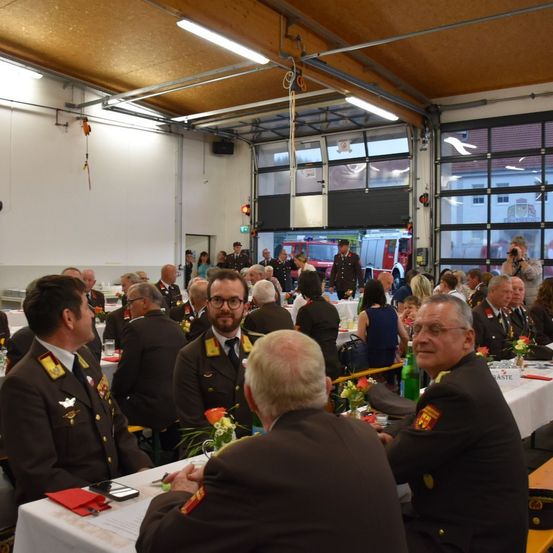 Eine große Gruppe von Männern und Frauen in Uniform sitzt an Tischen in einem Saal. Ein Mann mit Brille schaut einen anderen Mann an. Auf den Tischen stehen Blumen.