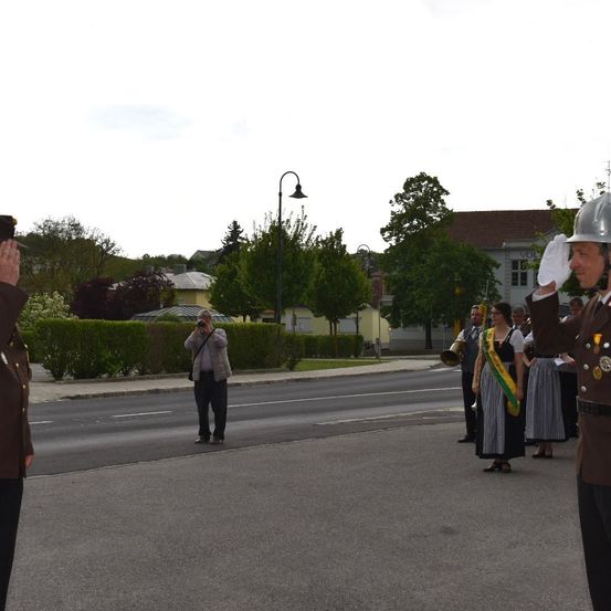 Eine Militärparade auf der Straße, bei der zwei uniformierte Offiziere salutieren. Dahinter hält eine Gruppe von Menschen in traditioneller Kleidung Trompeten. Im Hintergrund macht ein Fotograf das Ereignis fest. Gebäude und Bäume säumen die Straße.