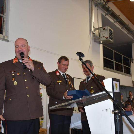 Ein Mann in Uniform spricht in ein Mikrofon an einem Podium, wobei zwei weitere uniformierte Männer hinter ihm stehen. Sie befinden sich in einem Saal mit einem Tisch und Stühlen im Hintergrund.