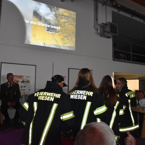 Eine Gruppe von Feuerwehrleuten steht in einem Raum. Eine Projektionsleinwand zeigt eine Feuerszene. Ein Mann in Uniform sitzt auf einer Bank links. Ein Tisch steht rechts.