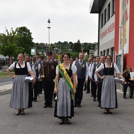 Eine Gruppe von Menschen in traditioneller Kleidung geht vor einem Gebäude, einige halten Musikinstrumente. Das Gebäude hat eine rote Wand mit weißen Schriftzügen.