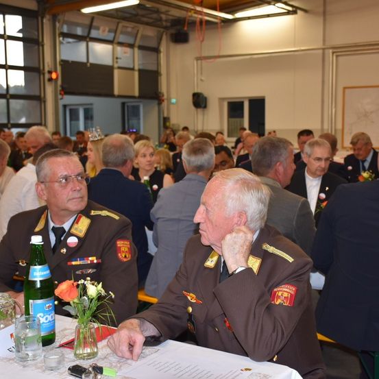 Ein Raum gefüllt mit Personen in Militäruniformen und Anzügen. Ein älterer Mann in Uniform sitzt an einem Tisch mit einer Blumenvase, einer Flasche und einem Menü. Andere Menschen sitzen und stehen hinter ihm.