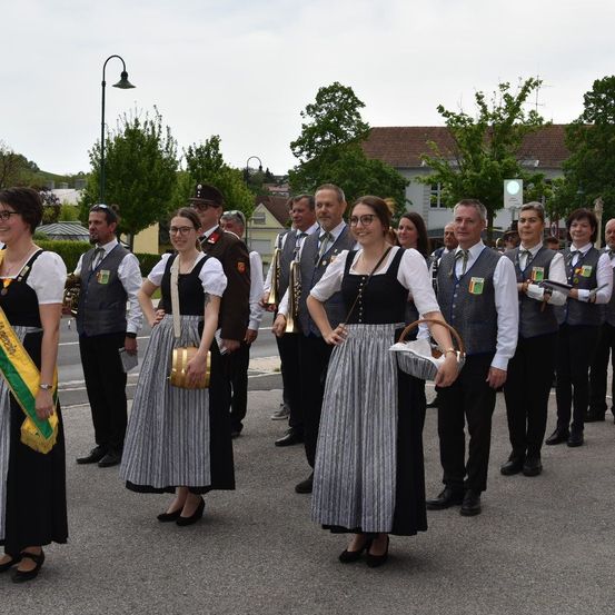 Eine Gruppe von Menschen in traditioneller Kleidung, einschließlich Musikinstrumente, steht auf einer Straße. Bäume und Gebäude sind im Hintergrund.