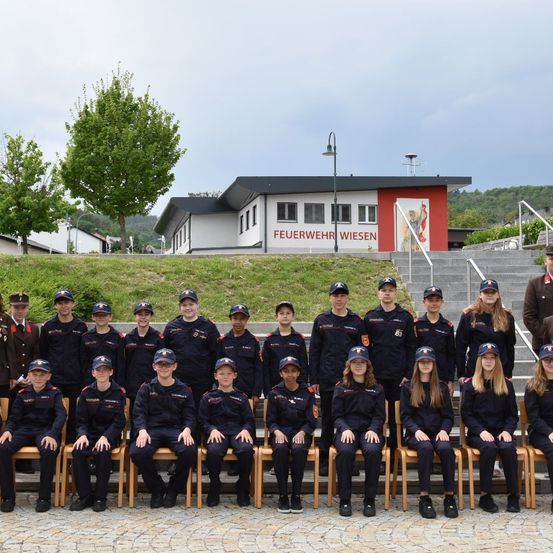Eine Gruppe junger Personen in Uniform posiert für ein Foto vor einem Gebäude mit der Aufschrift Feuerwehr Wiesen. Sie sitzen auf Holzstühlen mit einem Grashang und Bäumen dahinter.